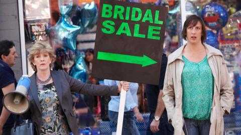 An older woman holds a megaphone in one hand and a sign in another. The sign reads 'BRIDAL SALE' and points at another woman, who looks fed up