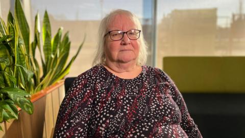 A woman with white hair just above her shoulders who is wearing black rectangular glasses. She is sat in a chair looking into the camera with a serious expression. She is wearing a spotty-striped dark red long-sleeved top. Behind her is a snake plant and a long green and black chair.