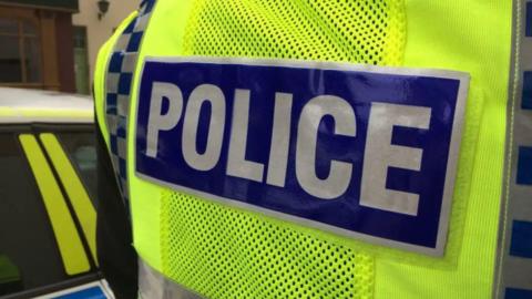 The back of a police officer wearing a bright yellow hi-vis jacket with "police" in capitals in silver and blue. The side of a police car is next to them.