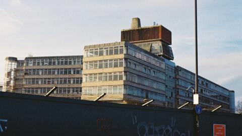 Sovereign House in Norwich is visible behind security hoardings, with its spiral glass staircase, concrete and glass offices and unique tower structure on top visible