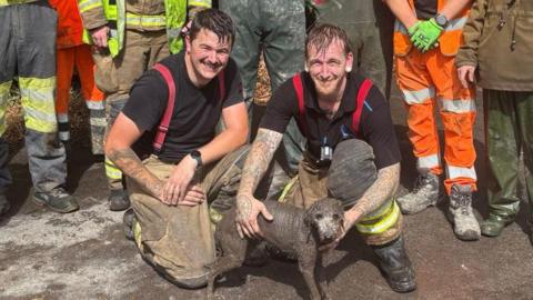 A group photo showing firefighters, council workers in hi-vis and local people in waders standing around a small dog who is covered in grey mud. They pose for a group photo on a gravel road and there is a white Iveco truck to their right