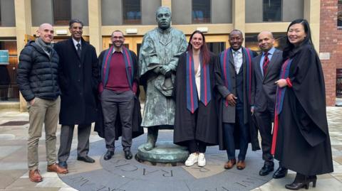 A posed group shot featuring students who took part in the specialist kidney training, together with consultants who created the court. They are five men and two women lining up either side of a metal statue outside Newcastle University.
Four of them, two men and two women, wear a graduation gown with a blue and red and blue stole. The other three men are dressed in formal wear.