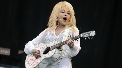 Dolly Parton performing on stage with a white guitar and wearing a white suit