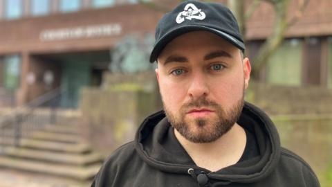 Simon Cobb has brown stubble and blue eyes. He is wearing a black hoodie and cap. Behind him is Portsmouth Family Court.