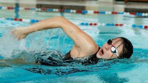 A young woman wearing goggles and a black swimming costume swims in an indoor swimming pool. She appears to be doing front crawl.