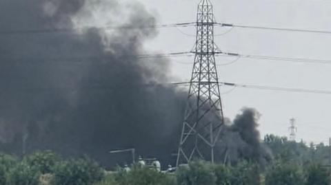 Thick black smoke is billowing into the sky close to a pylon. There are bushes in the foreground.