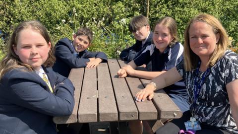 A number of pupils and one member of staff are sat around a wooden bench, all looking at the camera. One one side of the bench, a boy and a girl are sat. They are both wearing navy blue school blazers. On the other side of the bench a member of staff is wearing a black and white patterned top with two pupils sat next to her. One is a girl wearing a navy and white PE top and the other is a boy wearing black framed glasses and a navy blazer. 