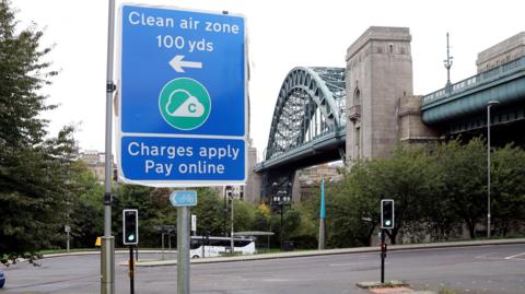 A view of a blue 'Clean air zone' sign in Newcastle city centre. It says 'Charges apply pay online'. Traffic lights on green can be seen behind the sign and a large metal bridge in the background.