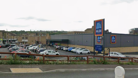 A google streetview image showing an Aldi supermarket. It looks like a small dark-grey warehouse. The car park outside is full.