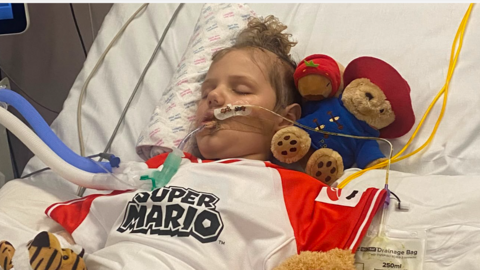 Little boy with tubes in his nose and arms on hospital bed and eyes closed surrounded by teddy bears