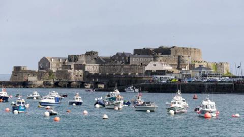 Castle Cornet, a large island castle on Guernsey that is largely surrounded by water. In the image there are several boats moored in front of the castle. There is also a full car park to the right of the structure. The day is largely clear with some light cloud coverage.