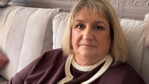 Sara Stead, woman with blonde hair dressed in a brown cardigan, sitting on a cream coloured couch.