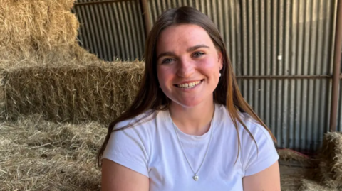 A young woman with long straight brown hair and blue eyes smiles at the camera. She is wearing a white T-shirt and a thin silver necklace with a small pendant on it. Behind her are bales of straw and a corrugated iron wall
