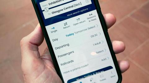 Hand holding a smartphone displaying the ScotRail app journey planner. The screen shows a search for train travel from Exhibition Centre (Glasgow) to Glasgow Central, with options for ticket type, travel date, departure time, passengers and railcards. The background is an out-of-focus tiled pavement.