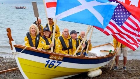A boat crew dressed in bright yellow wave a range of flags while sitting in a yellow, white and blue boat on the south of Scotland shoreline