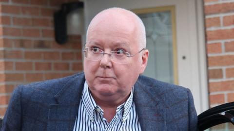 A bespectacled balding man in a blue sports jacket and striped shirt emerges from a car outside the front door of a modern house