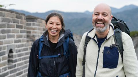 Contestants Yin and Gaz smile to the camera wearing outdoor jackets and backpacks