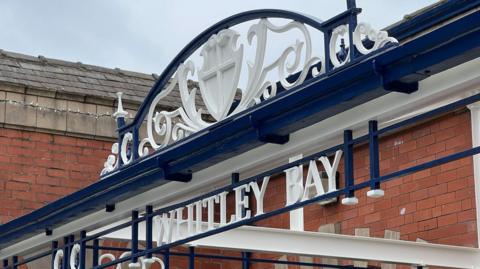 Newly painted Whitley Bay sign at the front of the station below a decorative archway. The sign is white and navy blue, with the wording in white. It is made of intricate metalwork and sits in front to the red brick station building, a small section of which is included in the image.