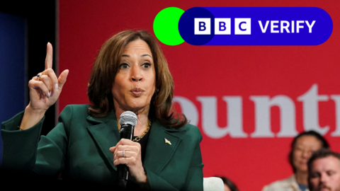 Kamala Harris speaking into a microphone. She's wearing  green jacket and pointing upwards with her right hand. The BBC Verify logo is in the top corner.