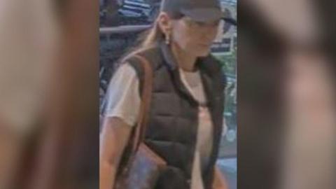 A woman with long hair and wearing a black cap, black gilet and white t-shirt. She is carrying a bag.