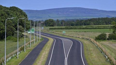 An aerial image of a section of the new Southern Link Road in Carlisle. The single carriage road has a cycle path on one side and is surrounded by green space. It snakes towards the fells in the background.