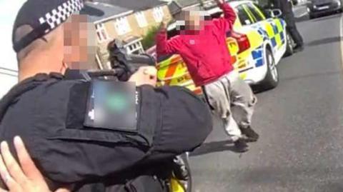 A police officer pictured from behind is wearing black uniform and aiming a gun at a man wearing a red jumper, who has his hands up. Behind this man is a police car. They are on a residential road.