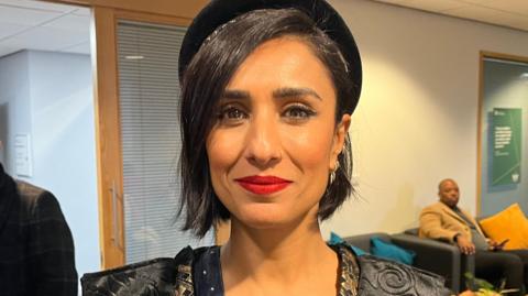 Anita Rani smiling and looking at the camera. She has medium length dark hair and is wearing a black hat. She is stood in a University of Bradford building, with seating and a door behind her. 