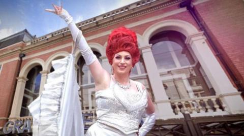 A woman with tall red hair is smiling at the camera standing outside a theatre. She is wearing a white dress and her right arm is outstretched above her head.