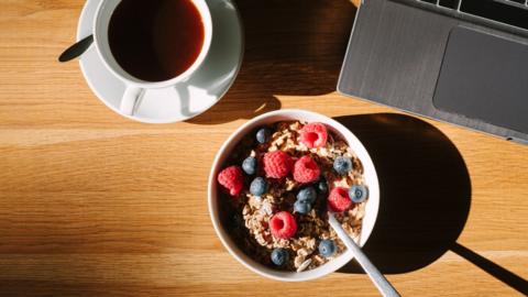 An aerial view of a bowl of granola with vibrant raspberries and blueberries, next to a laptop and a black coffee.