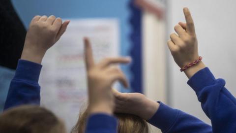 A close-up image of three children's arms raised in the air in a school classroom. They are all wearing blue jumpers.