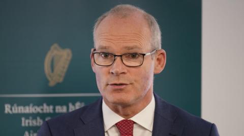 Simon Coveney standing in front of sign with an Irish government logo. He is wearing a suit and tie and glasses.