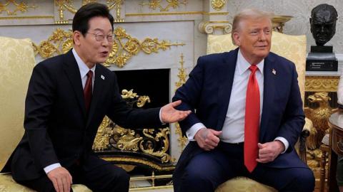 Lee Jae Myung and Trump sitting side by side in yellow armchairs in the Oval Office. They are wearing dark coloured suits with red ties. They're looking and smiling in the same direction.
