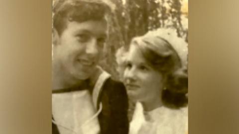 An old black and white photo shows a young man on the left, wearing what appears to be a naval uniform. On the right is a young woman with wavy fair hair, wearing a pale dress and a hat. He is looking at the camera and she is looking up at him.