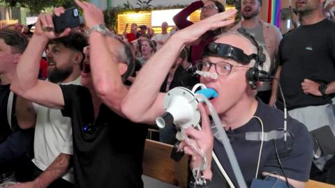 Prof Damian Bailey sits beside James Gallagher, who is wearing a head-mounted device with wires and sensors attached, as they watch football on TV in a packed pub