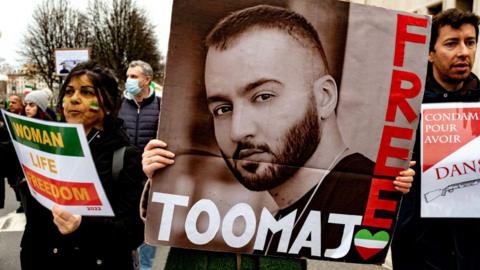 A large placard of Toomaj Salehi's face and the words 'free Toomaj' is held up at a protest. He is a young man with a beard, and the photo is in black and white. Two protestors stand either side of the placard.