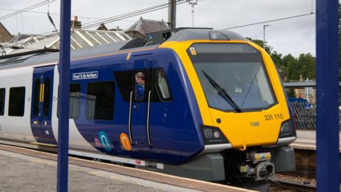 A Northern train which is painted in dark blue and yellow, in a station with the driver looking out of the window.