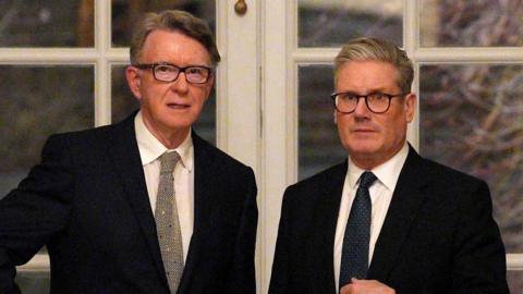 Peter Mandelson stands with Sir Keir Starmer in front of a set of double doors with white panes. Both men wear black suit jackets, white shirts and dark ties.