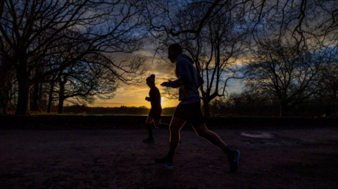 Silhoutted joggers run through a park. The sun is low in the sky, and there are trees surrounding the runners