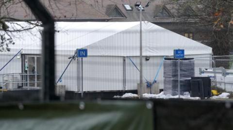 A large white tent behind a metal fence. The fence has blue signs reading "In" and "No entry" on it.