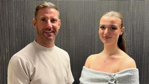 Chris (left) has short cropped brown hair and is wearing a beige t-shirt. Ava has her long brown hair tied up in a ponytail. She is wearing an off-the-shoulder grey top, nose ring and silver necklace.