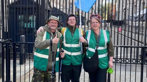 Rob, Lisa and son Drew outside gates in Downing Street. They are wearing dark green jackets and Rob Collins on the left is putting his thumb up.