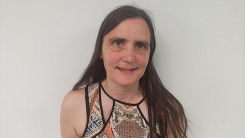 Alison Manning has long dark hair and is wearing a strappy dress with batik-type panels in brown and orange