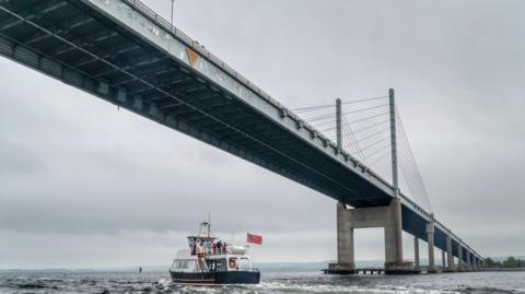 A dolphin-watching cruise boat sails under the bridge. The bridge is grey in colour, almost matching the colours of the sky above it and sea below.
