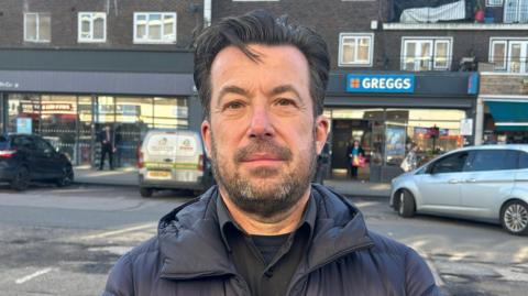 Simon Ford, Chair of the Harold Hill Shop Association, is standing in front of a road and a row of shops in Harold Hill, east London