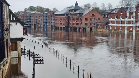 Flooded riverside area in York with water covering the walkway and reaching the ground floors of buildings. Several brick buildings, including converted warehouses and apartments, stand along the riverbank. Metal railings and benches are partially submerged.