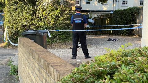 A police cordon at the side of a house with a community support officer, who has his back to camera, standing within it. The area cordoned off appears to be a gravel parking area.