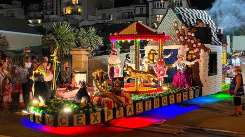 A brightly lit parade float decorated with flowers and lights, featuring a house, carousel, and costumed participants, travels along a street at night while spectators watch from the roadside.