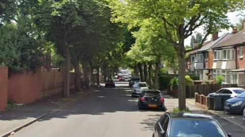 A tree-lined suburban street with mid-20th century houses visible down the right-hand side and brick walls and fencing along the left. Several cars are parked on the right side of the road. 