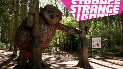A wooden troll in the forest and the Strange logo