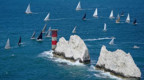 White outcrops with a red and white lighthouse, surrounded by blue water and racing sailing yachts